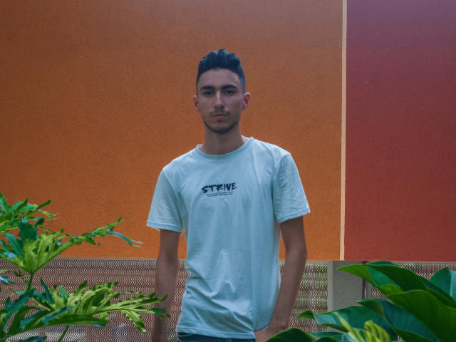 AlexOconor stands with hands in pockets, wearing a white t-shirt, short black hair, and a slight beard, in front of green plants. Orange wall in background.