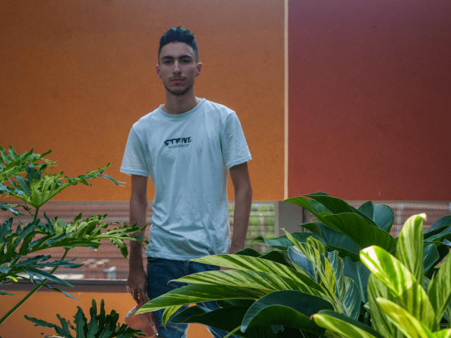 AlexOconor è in piedi di fronte a delle piante verdi, indossa una maglietta bianca e jeans blu, ha i capelli corti e scuri e un leggero sorriso, in un ambiente luminoso con pareti arancioni..