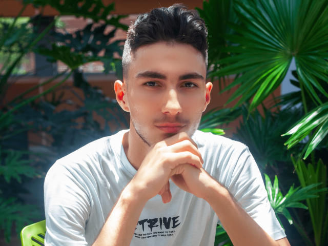 Alex Oconor mit kurzen, dunklen Haaren, grünen Augen und heller Haut, trägt ein weißes T-Shirt, die Hände sind vor dem Kinn gefasst, er ist von üppigen, grünen Pflanzen umgeben und blickt nachdenklich in die Kamera..