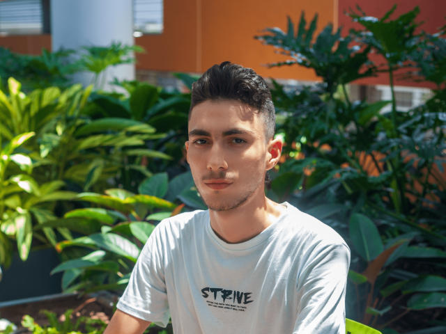 AlexOconor, avec des cheveux courts et foncés, une peau claire et une moustache, portant un t-shirt blanc, assis parmi des plantes vertes, regardant la caméra avec un léger sourire..