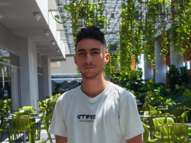 AlexOconor has short dark hair, a slight beard, and wears a white tee with "STONE" on it. Green plants hang overhead, and sun filters through. He looks calm.