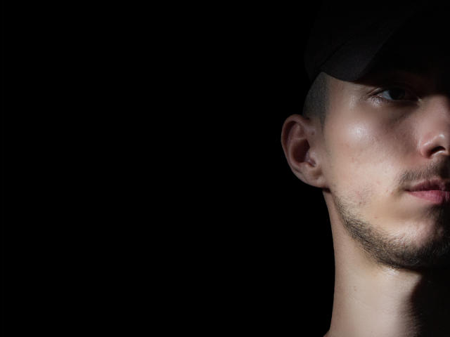 Il volto di AlexOconor, lato destro, con berretto nero, capelli corti e leggera barba, illuminato su sfondo scuro, che mostra una pelle luminosa e tratti del viso delicati..