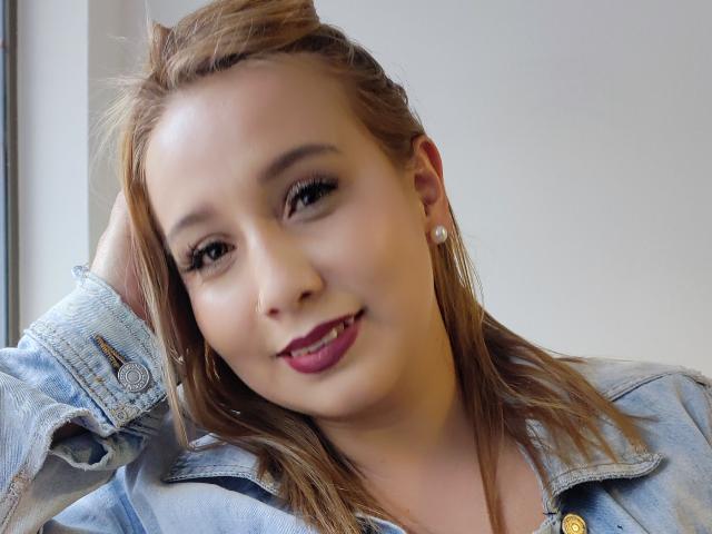Evelyn Swanth, con i capelli castano chiaro, una camicia di jeans blu, rossetto rosa e orecchini di perle, sorride con una mano tra i capelli, mostrando un viso carino e attraente..