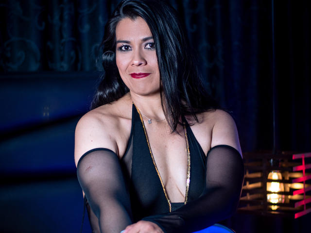 MartinaFox69 with black hair, red lipstick, and black sheer sleeves, wearing a deep cleavage black dress, smiling at the camera, arms crossed, looking sexy and confident.