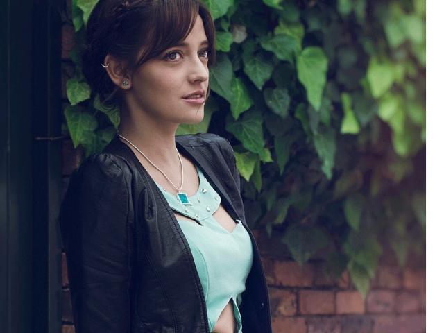 JaneRoa with brown hair, wearing a black leather jacket, light blue top, and necklace, standing against a brick wall with green ivy, has a cute face and small tits.