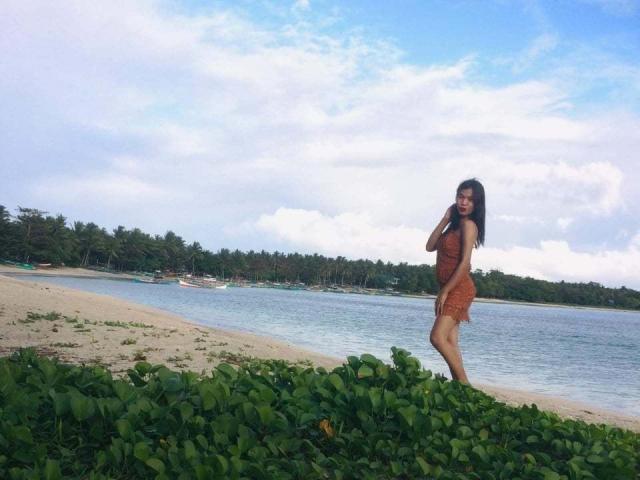 FantasyGodseyx è in piedi su una spiaggia, indossa un vestito arancione, con vegetazione verde in primo piano e uno sfondo di acqua blu calma, tiene in mano il telefono e sorride..