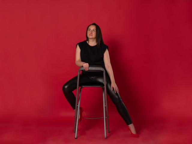 AbbyTaylorr with black shirt, black leather pants, and red background, sitting on metal chair with legs spread, hand on chair, looking slightly upward, barefoot.