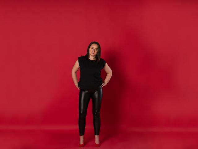 AbbyTaylorr stands with hands on hips, wearing a black top, black leather pants, and red heels, against a solid red background. She has a serious expression.