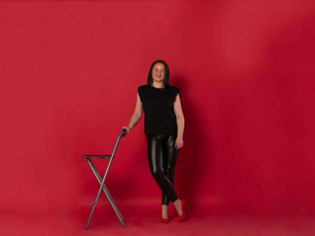 AbbyTaylorr leans on a metal chair, wearing a black top, shiny black leather pants, and red shoes, with a relaxed, confident smile, standing against a solid red background.