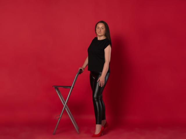AbbyTaylorr steht in einem schwarzen T-Shirt und glänzenden schwarzen Lederhosen, hält einen Metallstuhl und trägt rote High Heels, vor einem roten Hintergrund. Ihr Hintern ist leicht sichtbar..