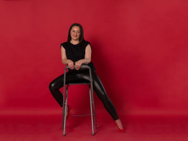 AbbyTaylorr sits on a metal chair with legs spread, wearing a black sleeveless top and shiny black leather pants, red shoes, smiling with straight brown hair.