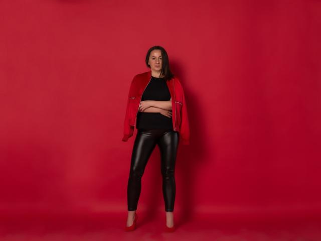 AbbyTaylorr stands with arms crossed, wearing a red jacket, black top, and shiny black leather pants, against a red background, with her brown hair down, and nude heels.