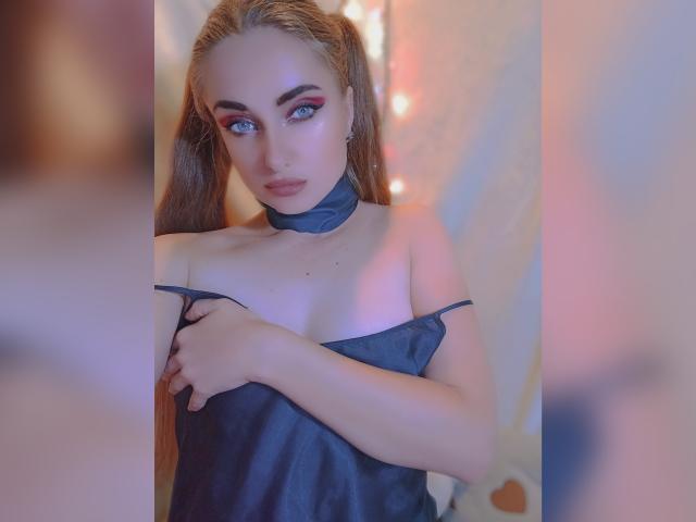 KariMillie with blue eyes, wearing a black satin top, has her hand on her tits, looking into the camera with pouty lips, blonde pigtails, and a black choker.