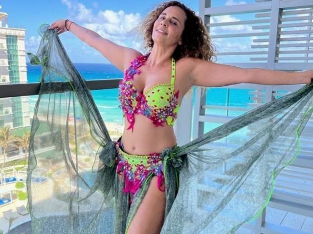 Shakirita with curly hair, wearing a neon green and pink floral bikini, holding a green sheer scarf, standing on a balcony with a blue ocean view.