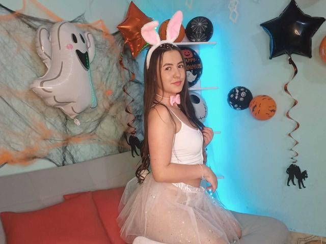 CamilaGomez with long brown hair, white bunny ears, pink bow, white tank top, and sheer pink tulle skirt, showing her ass, sitting on a bed, looking at camera.