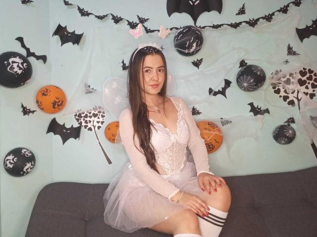 CamilaGomez with long brown hair, white lace dress, and fairy wings, sitting on a gray couch, wearing a headband with bat ears, and knee-high socks, in a Halloween setup.