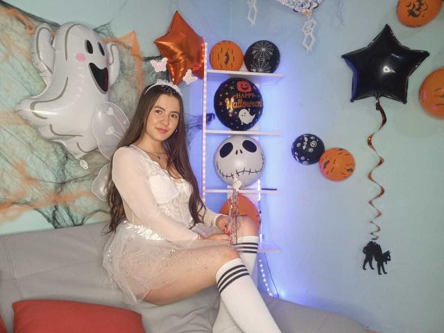 CamilaGomez with long brown hair, wearing a sheer white dress, sitting on a gray couch, white knee socks, surrounded by Halloween balloons and decorations, with a ghost balloon.