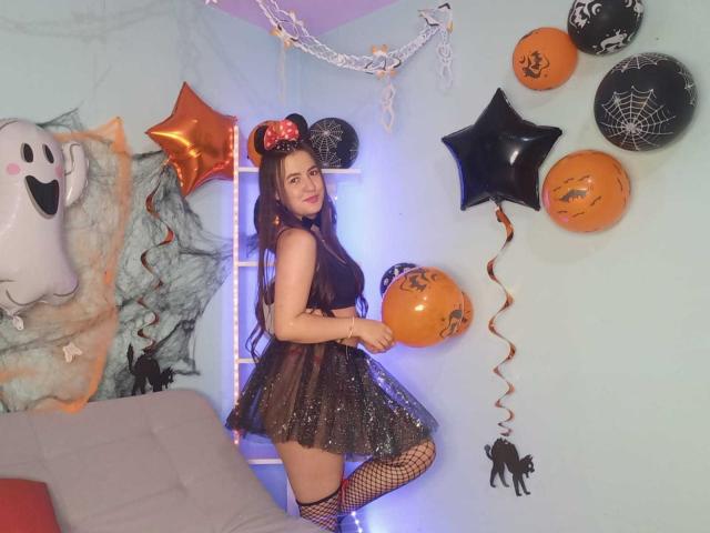 CamilaGomez stands in a Halloween-themed room, wearing a sparkly black mini dress, fishnet stockings, and a Mickey Mouse headband with orange bows, holding orange balloons.