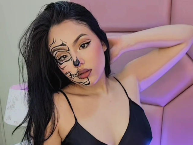 HaleiBeckham con rostro tatuado de negro, cabello largo y negro, top con tirantes negros, mano detrás de la cabeza, mirando a la cámara, iluminación morada y rosa. Pechos visibles, pequeños, lindos..