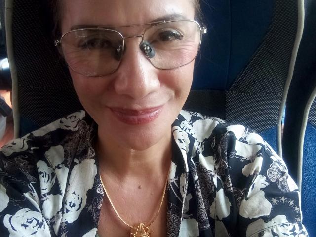 Dominique, une femme d'âge mûr, portant des lunettes, souriante, vêtue d'une chemise noire à motifs floraux et d'un collier en or, assise sur un siège de voiture bleu. Elle a un joli visage et une petite tache de naissance..