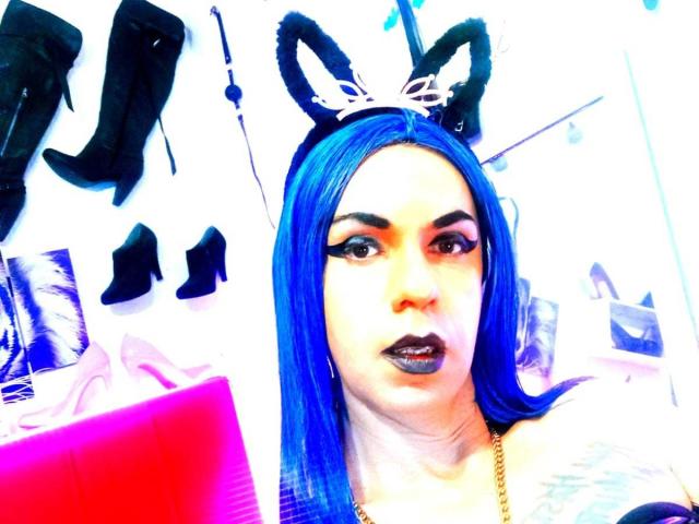 Bellanida with blue hair, black cat ears, and black lipstick, wearing black high heels in the background. She's looking at a pink object with a confident expression.