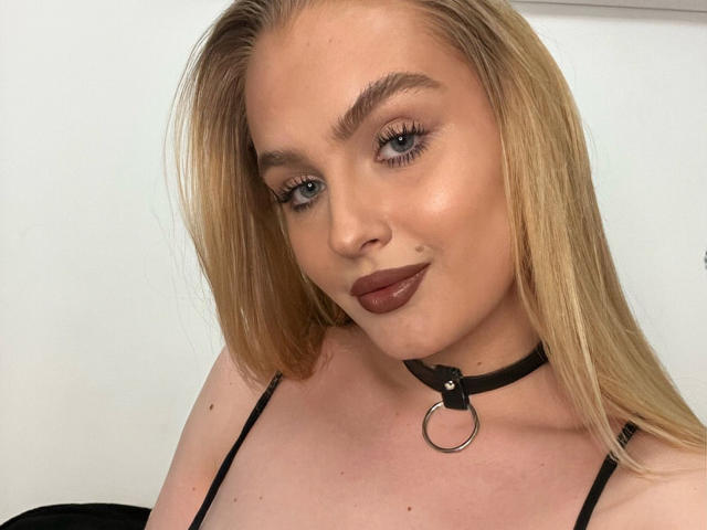 Olivia: Een vrouw met blond haar, blauwe ogen en donkere lipstick, die een zwarte choker met een metalen ring draagt en de camera aankijkt met een lichte glimlach. Haar borsten zijn zichtbaar..