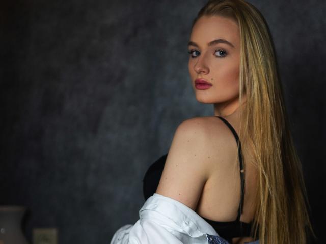 Olivia, les cheveux longs et blonds, en soutien-gorge noir et chemise blanche ouverte, regarde par-dessus son épaule, montrant son dos nu et ses petites seins mignons. Elle a les yeux bleus et les lèvres roses..