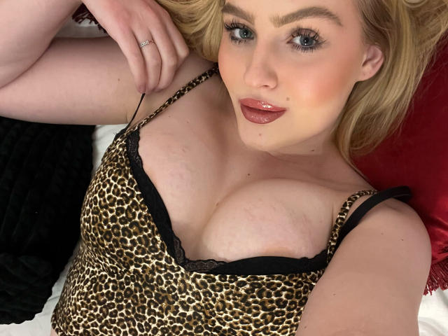 Olivia, les cheveux blonds, les yeux bleus et le rouge à lèvres rose, allongée, portant un haut à léopard avec de la dentelle noire, montrant ses seins volumineux, ronds et pâles..
