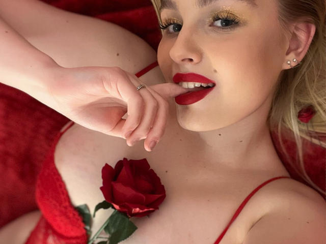 OliviaBent with red lipstick, biting her finger, wearing red lace, a rose between her tits, and a red rose in her hair, on a red blanket.