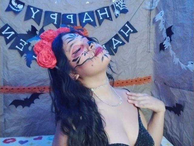 Rossy Granger con trucco nero, un fiore rosso tra i capelli, un abito nero e una collana a catena, con la testa inclinata e una mano sul petto, che mostra il seno, in una foto a tema Halloween..