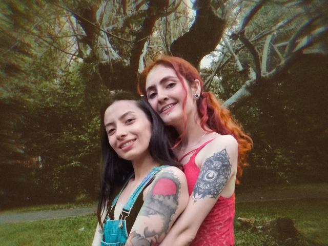 BethAndRoseI, con cabello rojo y tatuajes, con los brazos alrededor de BethAndRoseI de origen asiático, con cabello negro, sonriendo. Se puede ver el vestido rojo de BethAndRoseI, sus tatuajes y los aretes. El entorno es al aire libre..