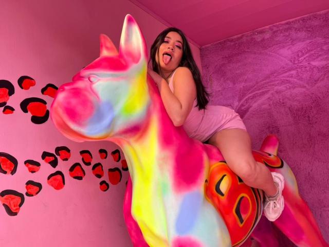 Veronica, mit langen braunen Haaren, einem pinken Tanktop und pinken Shorts, sitzt auf einem farbenfrohen, regenbogenfarbenen Pferd, steckt die Zunge heraus, trägt weiße Turnschuhe und befindet sich in einem pinken Raum..