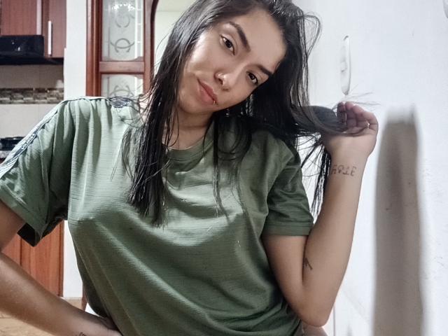 SharonKlart with long black hair, green and black shirt, bent over, showing her large, tattooed ass in pink panties, looking back with a slight smile.