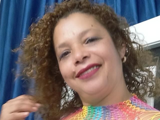 Victoria Borunys, con i capelli castani ricci, sorridente, con rossetto rosa e una maglietta a motivi arcobaleno, tiene i capelli, sfondo con tenda blu. Il suo seno non è visibile..