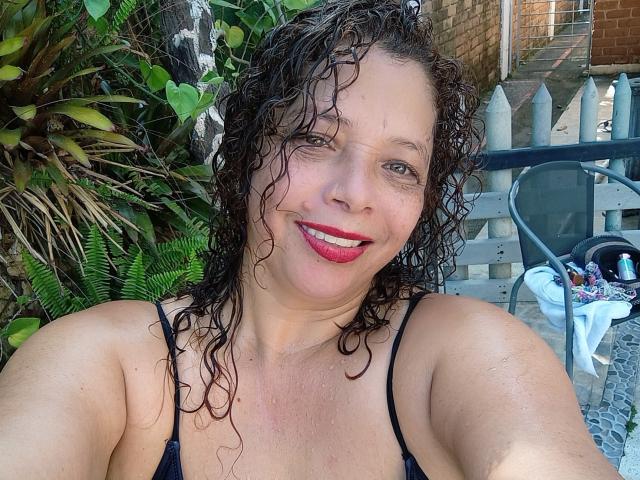 Victoria Borunys, con cabello oscuro y rizado, labios rojos, sonriendo a la cámara, vestida con una blusa negra de tirantes finos, mostrando sus senos, sentada afuera en un día soleado..