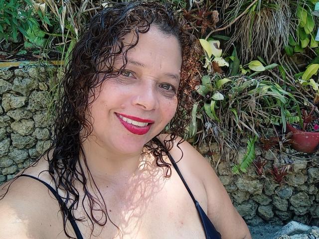 VictoriaBorunys with curly, wet brown hair, smiling, wearing a black spaghetti strap top, showing her tits, pink lipstick, and fair skin. Background is green foliage.