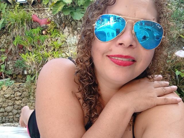 VictoriaBorunys with curly brown hair, red lipstick, and blue reflective sunglasses, smiling, arms crossed, wearing a black top, outdoors, showing her boobs, sitting on a white surface.