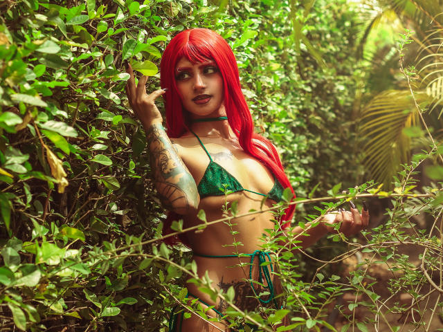 SerenaDeStark com cabelo ruivo, biquíni verde, tatuagens e um colar, em meio a uma vegetação densa, tocando as folhas com um sorriso sedutor, mostrando seus seios e um pouco de sua bunda..