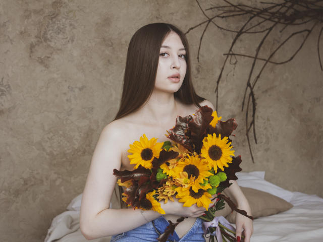 EllyCat with long brown hair, holding sunflowers and dark brown leaves, topless, showing small tits, and wearing blue jeans, against a textured wall.