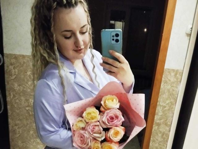 Jude Valenti, com cabelos loiros cacheados, vestindo uma camisa roxa clara, segurando um buquê de rosas rosa e amarelas, tirando uma selfie no espelho com um telefone azul..