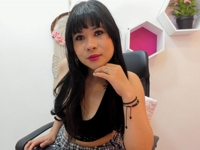 LourrieAnn mit schwarzen Haaren, rotem Lippenstift, trägt ein schwarzes Oberteil, das ihre kleinen Brüste zeigt, einen karierten Rock, einen schwarzen Stuhl, einen Ring am Finger, Armbänder und im Hintergrund steht eine Topfpflanze..