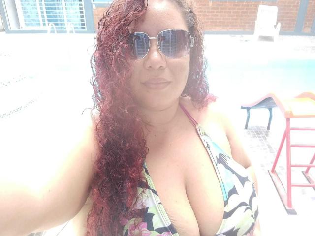 AdelineLox tem cabelos ruivos cacheados, está usando óculos de sol e um biquíni floral que mostra seus seios grandes e arredondados. Ela está sentada perto de uma piscina, com um fundo claro e ensolarado..