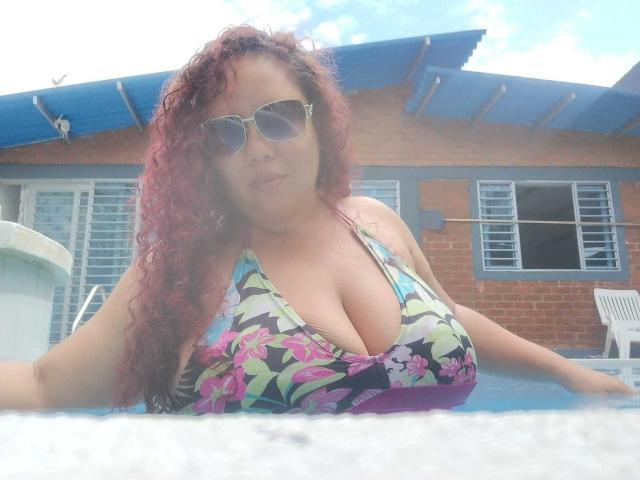 AdelineLox, com cabelos ruivos cacheados, usando óculos de sol, um biquíni colorido com estampas florais, mostrando seios grandes, flutuando em uma piscina, com as mãos apoiadas na borda da piscina..