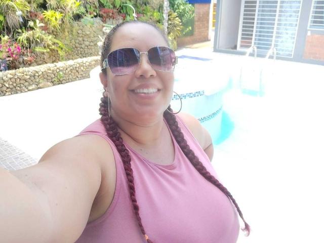 Adeline Lox, uma mulher corpulenta com cabelo ruivo trançado, vestindo uma regata rosa e óculos de sol, sorrindo para a câmera perto de uma piscina azul brilhante. Seus seios estão visíveis..