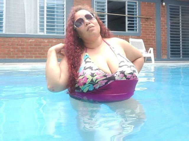 AdelineLox con i capelli rossi e ricci, grandi seni, indossa un bikini colorato con motivi floreali e viola, occhiali da sole, in piedi in una piscina blu, mani vicino alla testa..