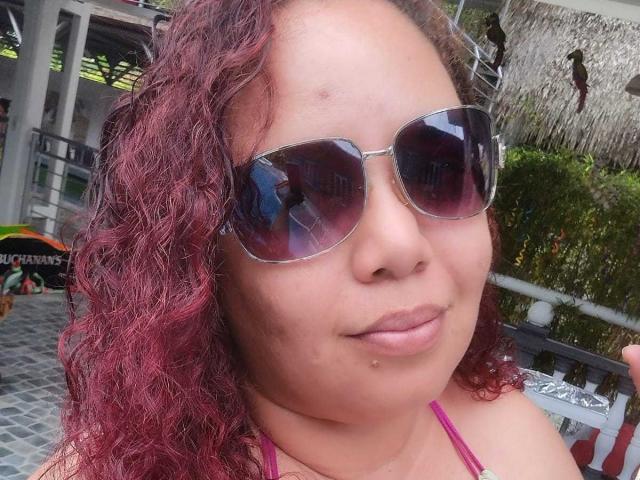 Adeline Lox, con cabello rojo rizado, lleva gafas de sol tipo aviador oscuras, tiene una pequeña peca en la mejilla derecha y un piercing en el labio, y está sonriendo ligeramente..