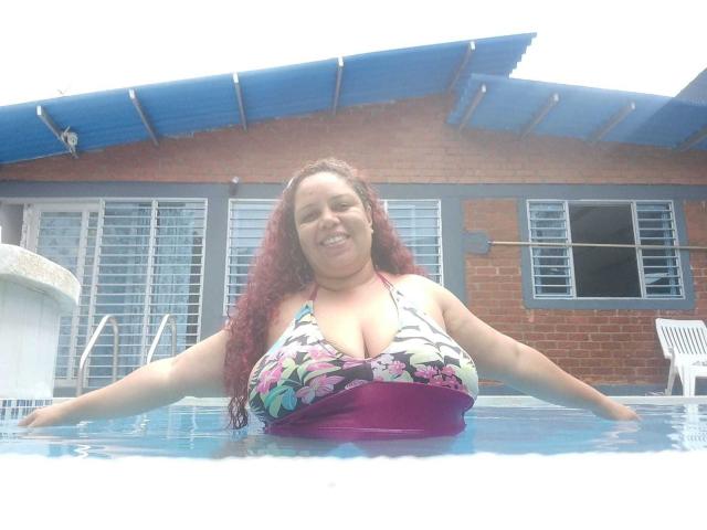 AdelineLox, uma mulher plus size com cabelos ruivos e cacheados, está em uma piscina, sorrindo com os braços estendidos, vestindo um biquíni floral que mostra seus seios grandes e proeminentes..