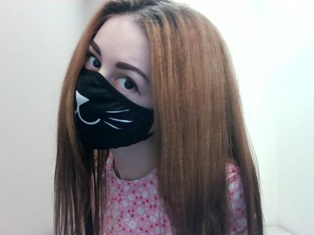 EvaRouseX mit langen, braunen Haaren, die eine schwarze Katzenmaske trägt, die ihren Mund bedeckt, und ein pinkes, geblümtes Hemd, hat große, braune Augen, die aufblicken und einen niedlichen Ausdruck zeigen..