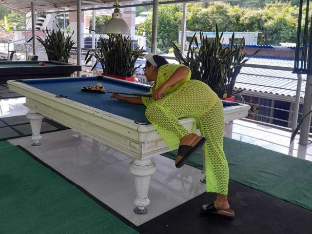 Mara Roberts, con un vestido de malla de color amarillo neón, inclinada sobre una mesa de billar blanca, mostrando su trasero, con una pierna levantada, usando chanclas y un gorro blanco..