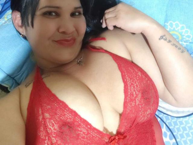 LadyMaturee, con senos grandes y voluptuosos, vestida con un sujetador de encaje rojo, acostada sobre una toalla azul, sonriendo a la cámara, cabello negro, tatuajes en el brazo, lápiz labial rosa..